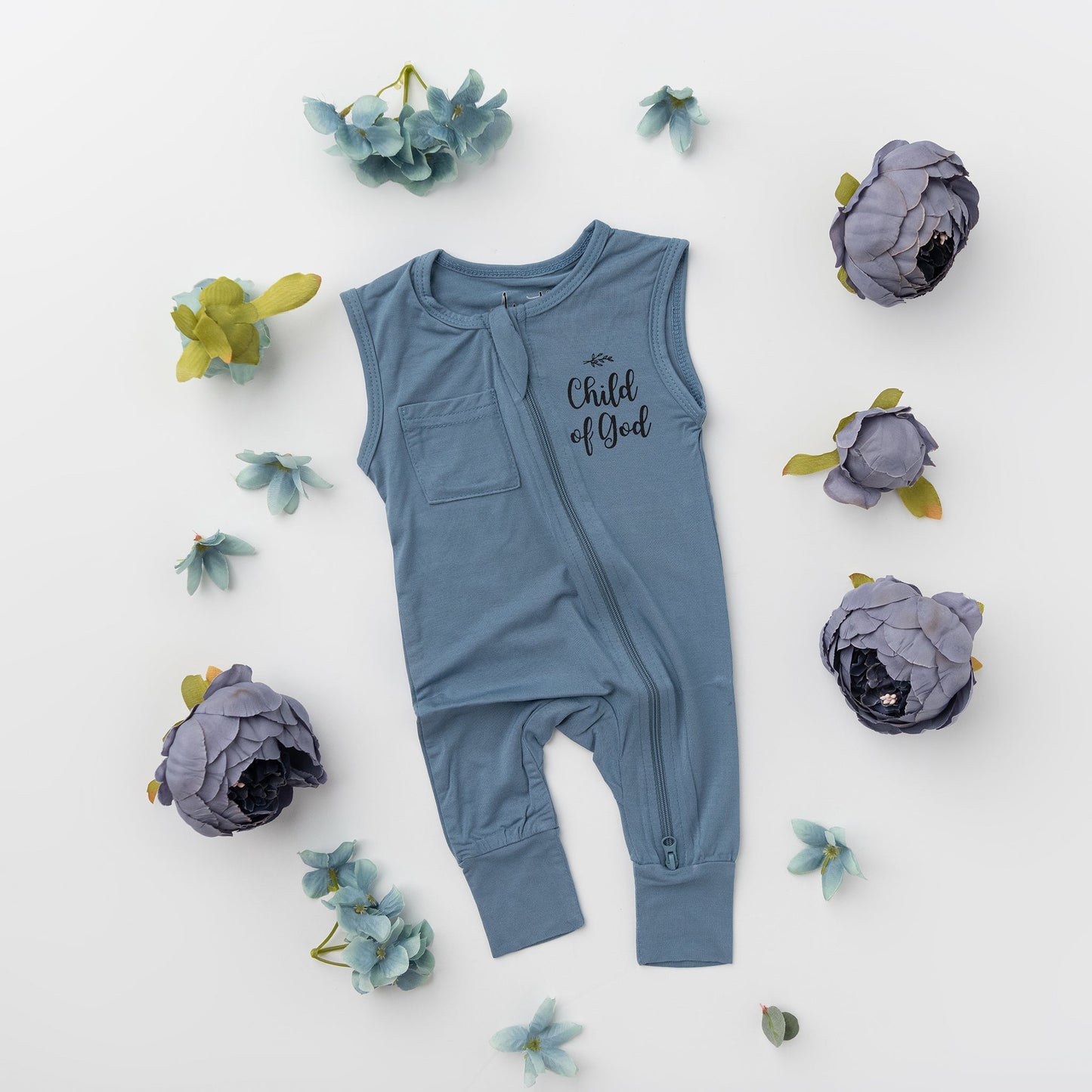 A flat lay of the Apostle Living Inc. blue "Child of God" romper with a zipper and chest pocket, surrounded by various blue flowers and leaves.