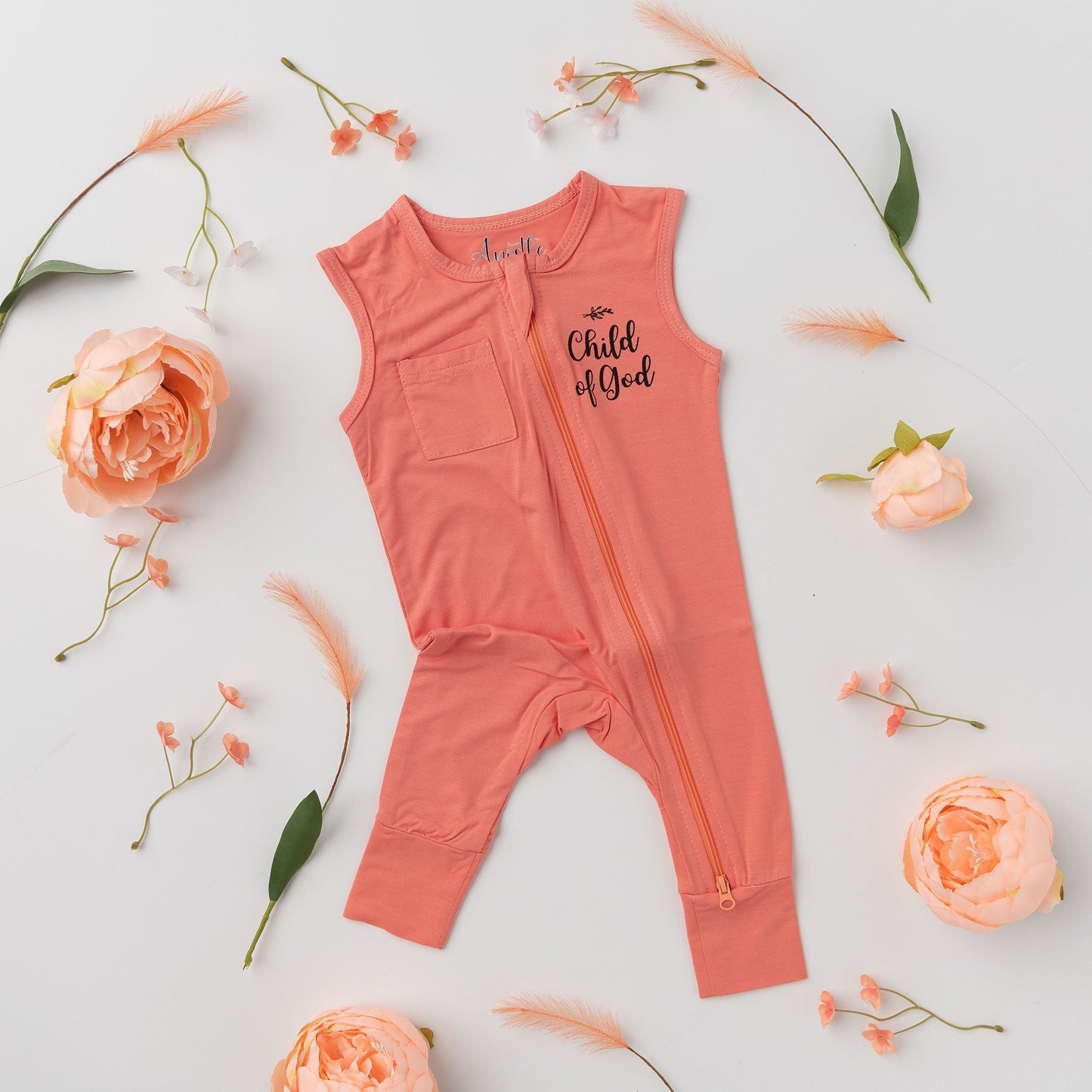 A flat lay of the Apostle Living Inc. pink "Child of God" romper with a zipper and chest pocket, surrounded by small flowers and feathers.
