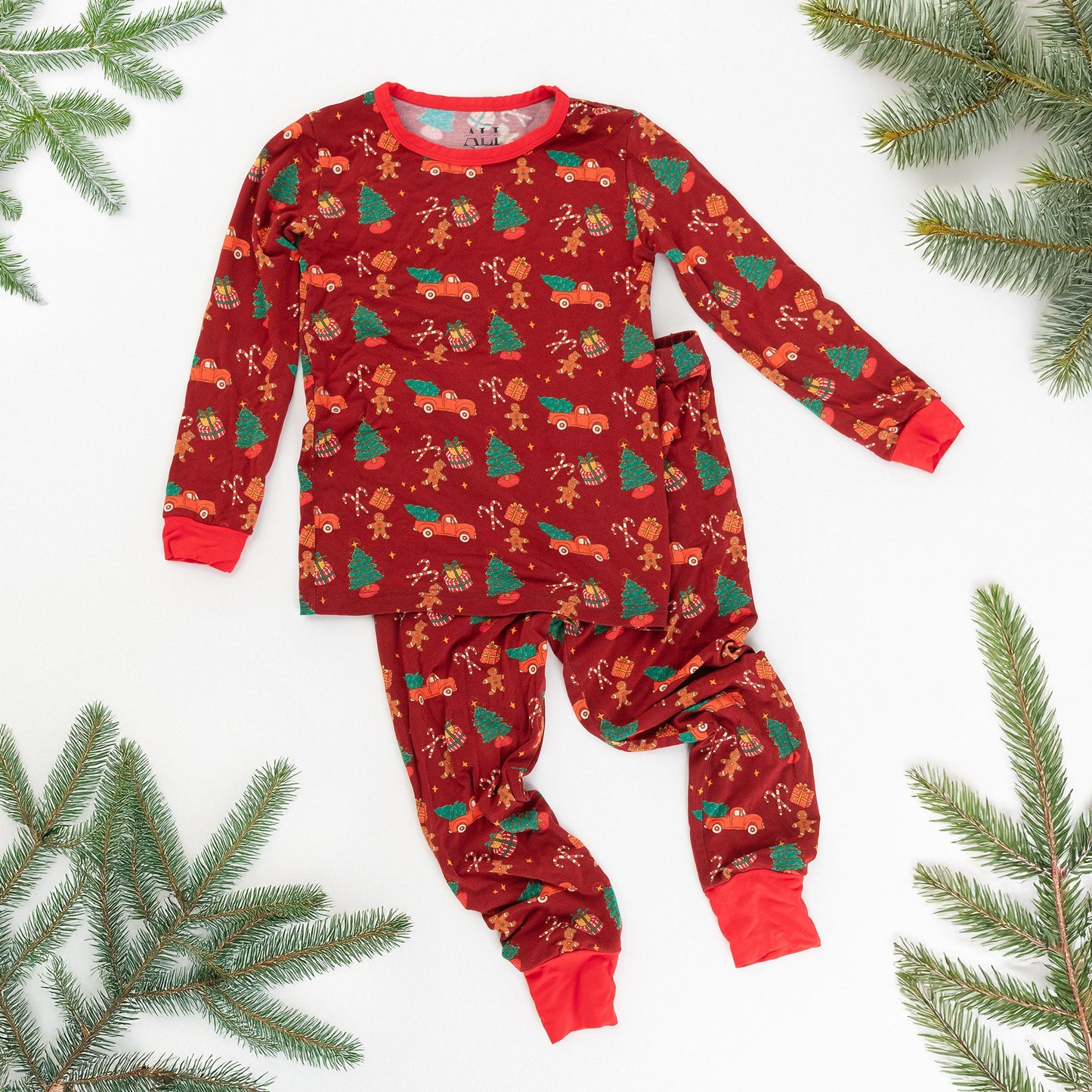 A flat-lay product shot of a red children's pajama set in soft bamboo fabric with a festive Christmas print, framed by green pine sprigs.