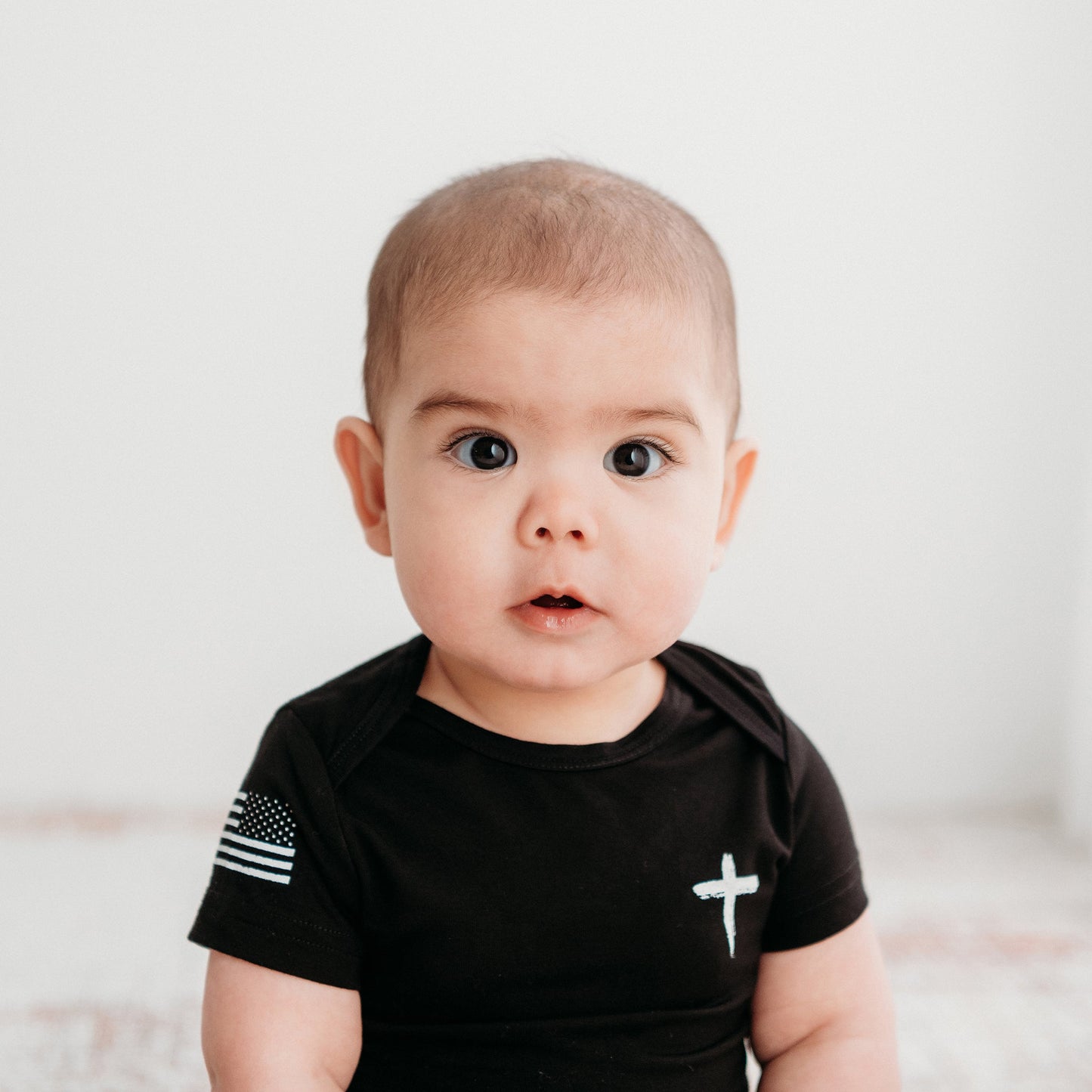 Close-up of a baby wearing the black Apostle Living Inc. Faith and Freedom onesie, which features a white cross and an American flag with a thin blue line on the sleeve.
