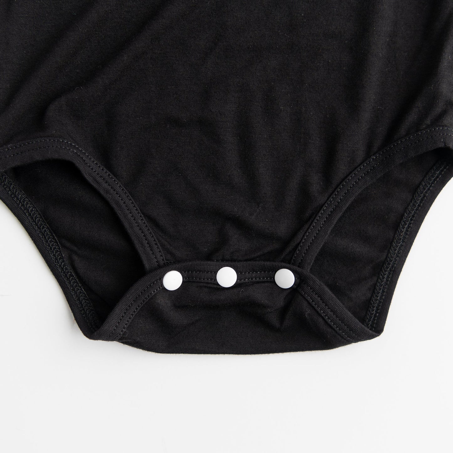 A close-up detail shot of the three-snap closure on the bottom of the Apostle Living Inc. Faith and Freedom baby onesie in black, showing the three white snaps.