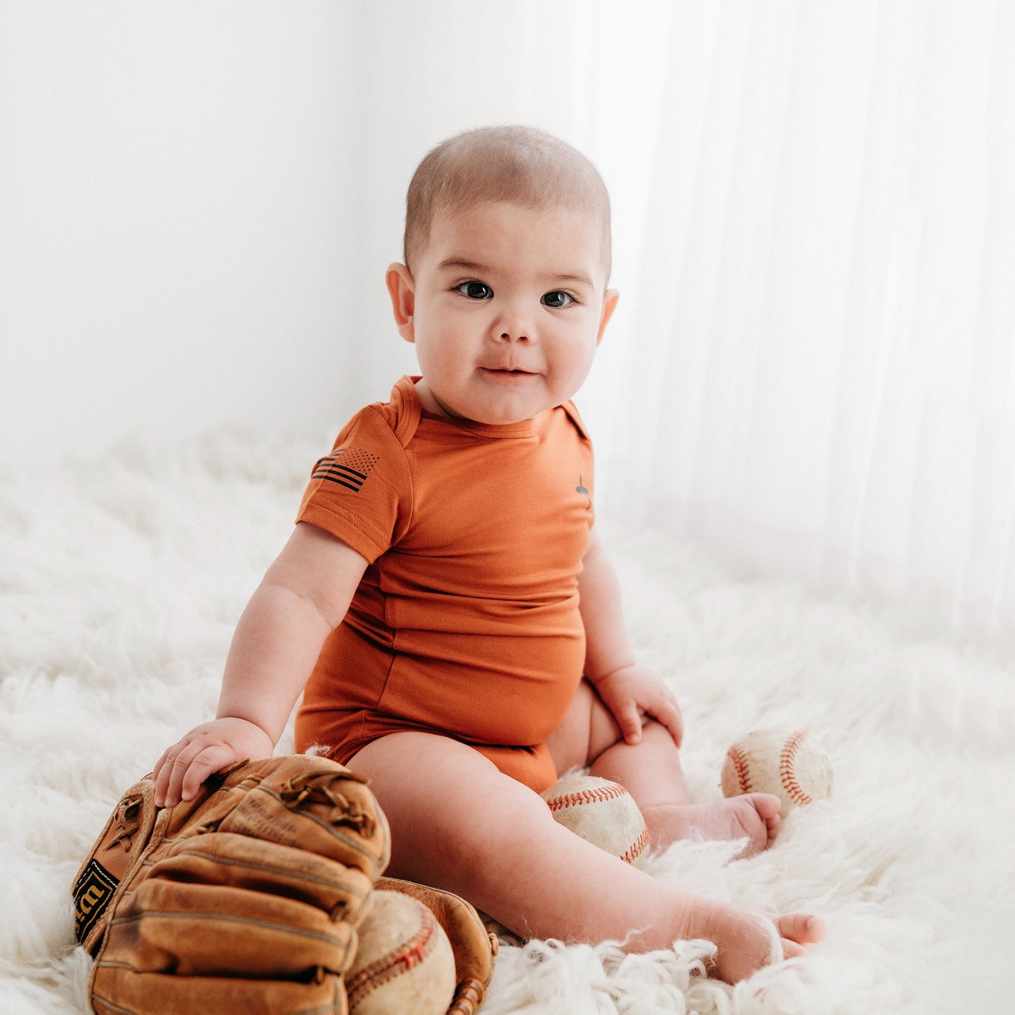 A baby from Apostle Living sits on a white fuzzy rug, wearing a rust-colored "Faith and Freedom" onesie with a small distressed U.S. flag graphic on the sleeve, surrounded by a baseball glove and baseballs.