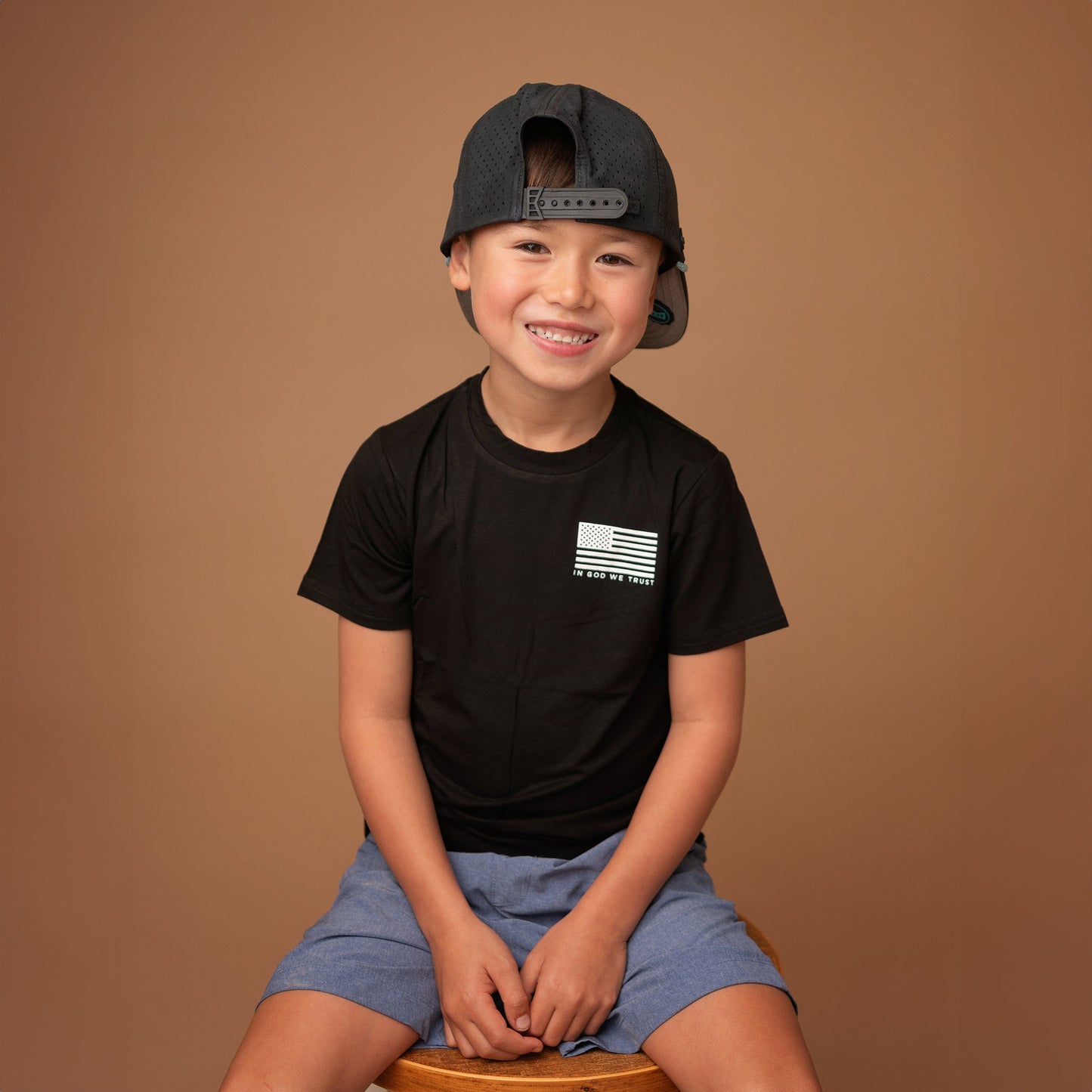 A young boy from Apostle Living sits barefoot on a wooden stool, smiling and wearing a black patriotic tee with a small distressed U.S. flag graphic and the text 'IN GOD WE TRUST' on the chest.