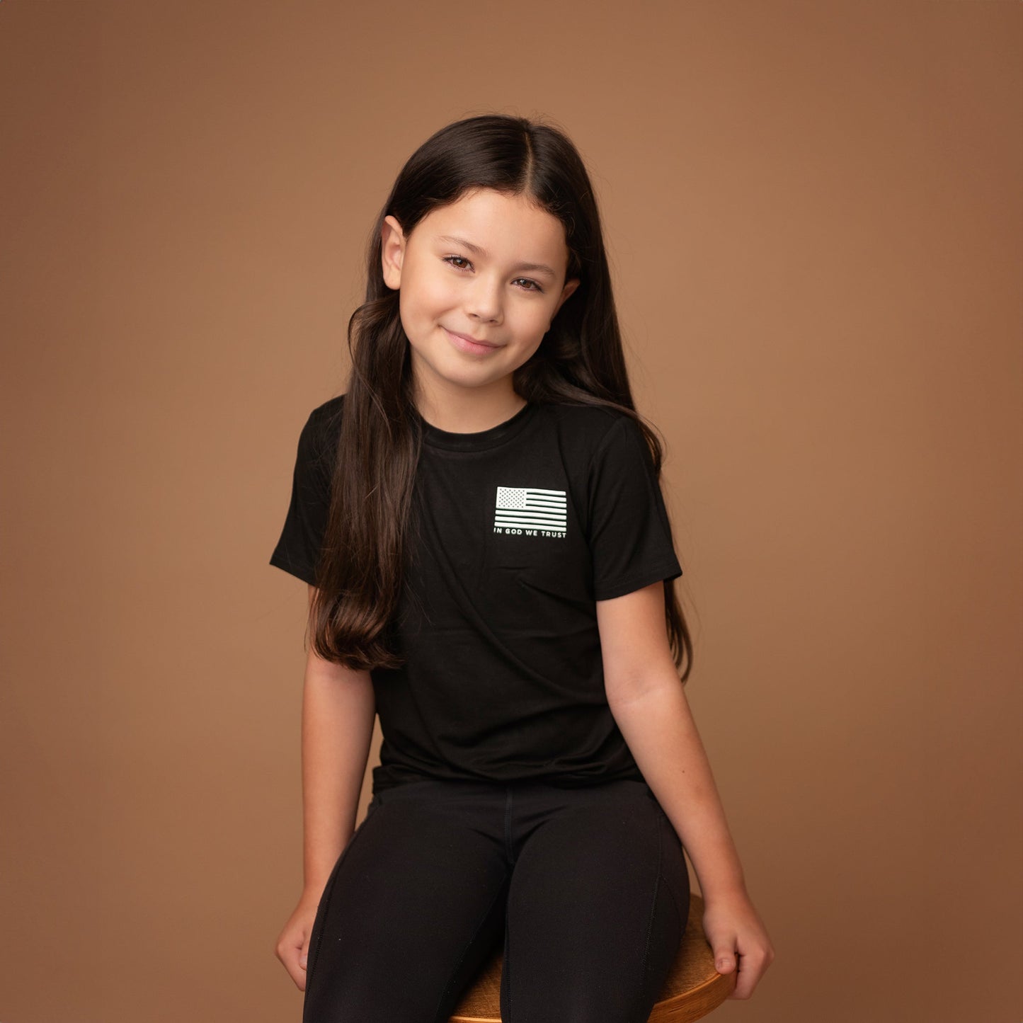 `A young girl from Apostle Living sits on a wooden stool, wearing a black patriotic tee with a white distressed USA.