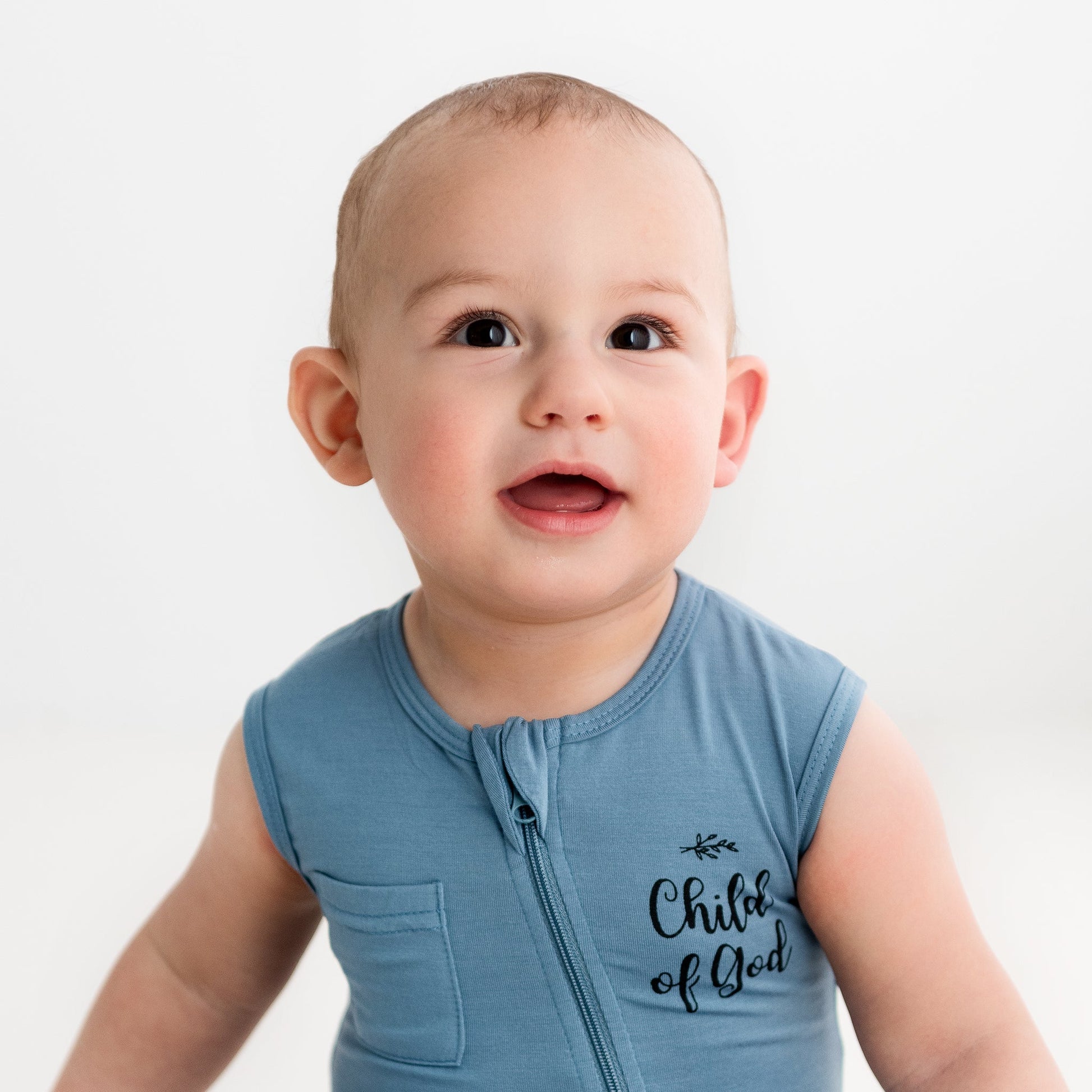 A close-up of a baby smiling while wearing the Apostle Living Inc. blue "Child of God" romper with a zipper and chest pocket.
