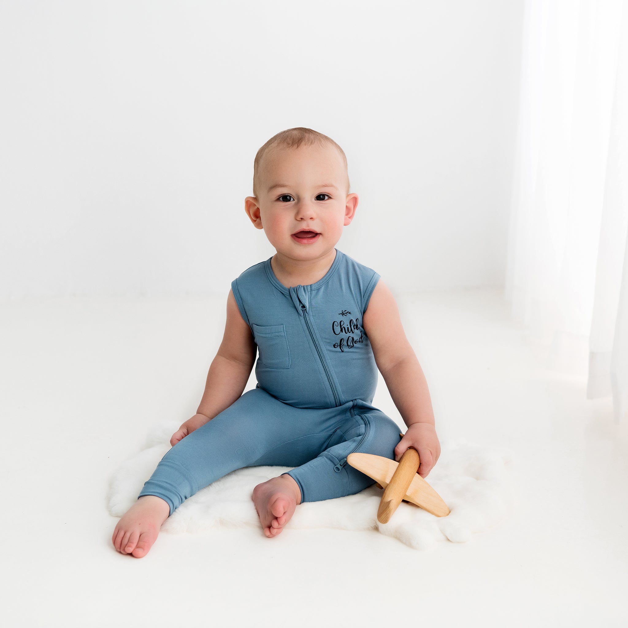 An infant sits on a fluffy white rug, smiling and holding a wooden toy airplane while wearing the Apostle Living Inc. blue 