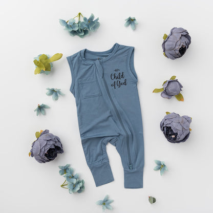 A flat lay of the Apostle Living Inc. blue "Child of God" romper with a zipper and chest pocket, surrounded by various blue flowers and leaves.