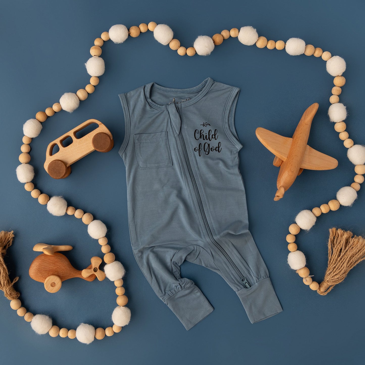 The Apostle Living Inc. blue "Child of God" romper is shown in a flat lay with a beaded garland and wooden toys, including a bus, plane, and helicopter.