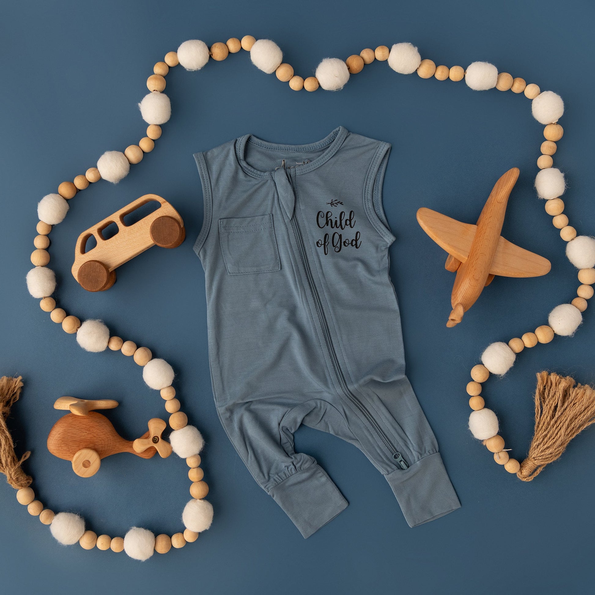 The Apostle Living Inc. blue "Child of God" romper is shown in a flat lay with a beaded garland and wooden toys, including a bus, plane, and helicopter.