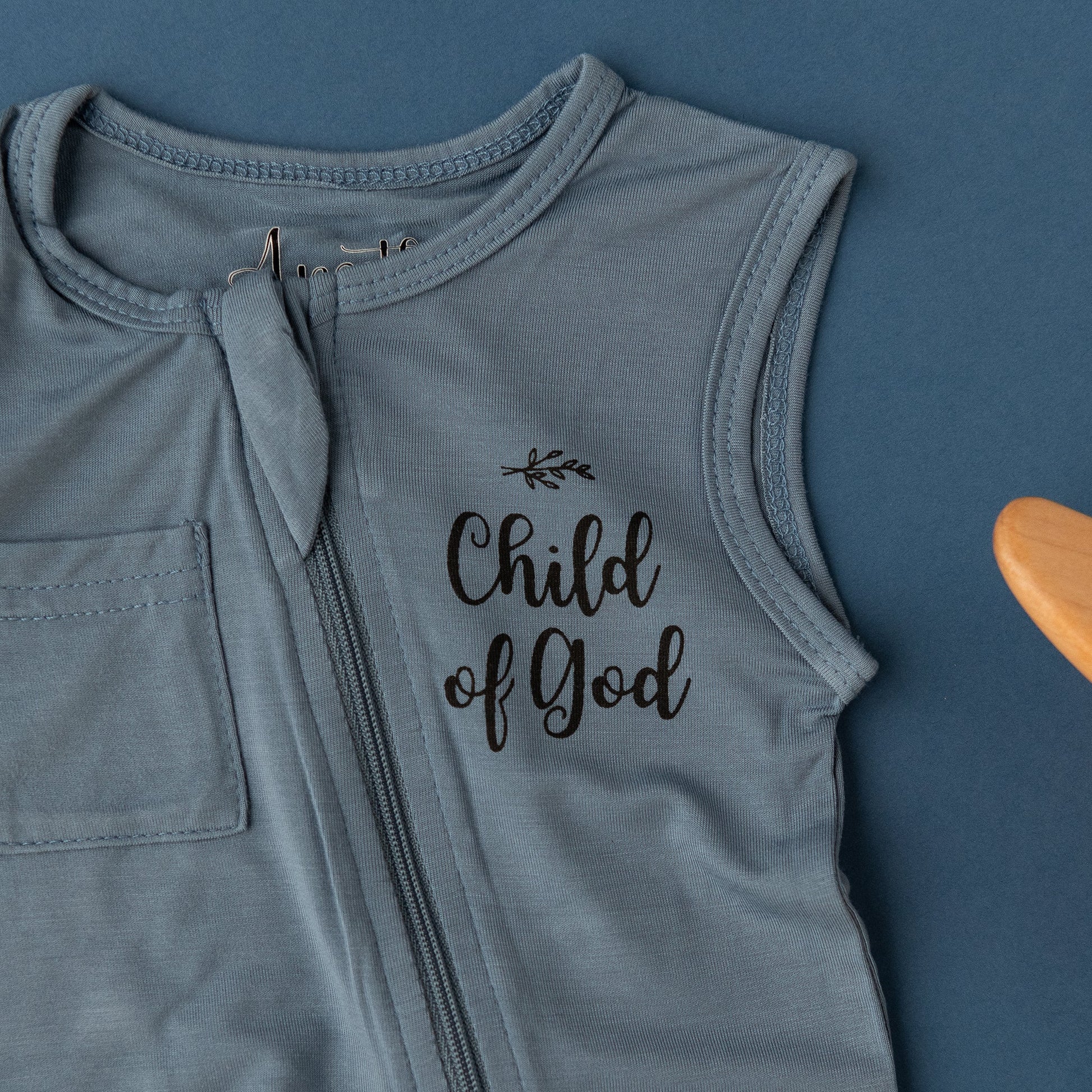 A close-up detail shot of the Apostle Living Inc. blue "Child of God" romper with a chest pocket and zipper, laid flat on a blue background.