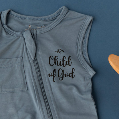 A close-up detail shot of the Apostle Living Inc. blue "Child of God" romper with a chest pocket and zipper, laid flat on a blue background.