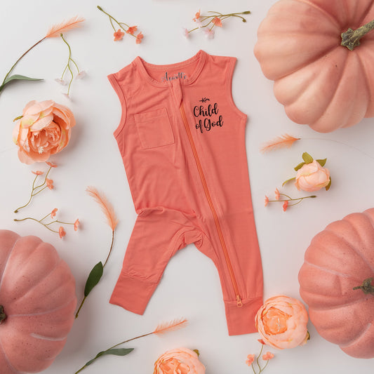 An Apostle Living Inc. pink "Child of God" romper with a full-body zipper, styled in a flat lay with small flowers, feathers, and pink pumpkins.