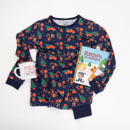 A flat-lay product shot of a navy blue children's Christmas pajama set from Apostle Living, made of soft bamboo fabric and styled with a hot cocoa mug and a Rudolph the Red-Nosed Reindeer book.