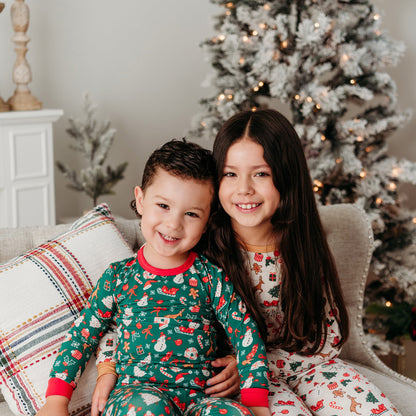 Two children in Christmas-themed bamboo pajamas sitting on a couch with a decorated Christmas tree in the background.