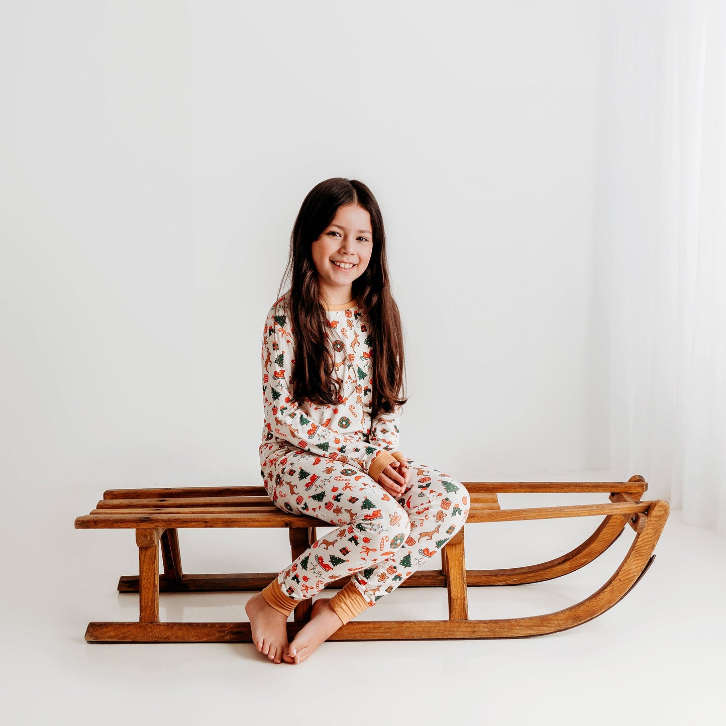 A young girl sits on a vintage wooden sled, smiling and wearing a soft ivory bamboo Christmas pajama set with a festive print.