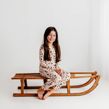 A young girl sits on a vintage wooden sled, smiling and wearing a soft ivory bamboo Christmas pajama set with a festive print.