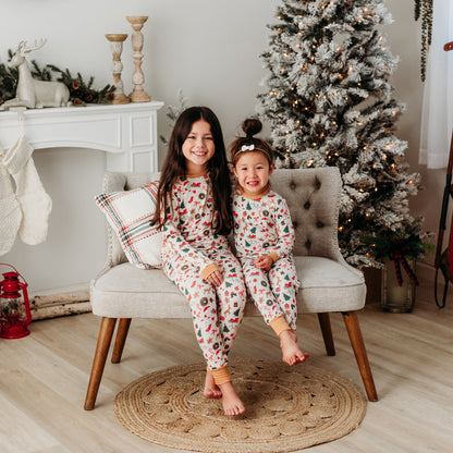 Two girls smiling and sitting on a bench in a holiday-decorated room, wearing matching soft ivory-colored bamboo Christmas pajamas.