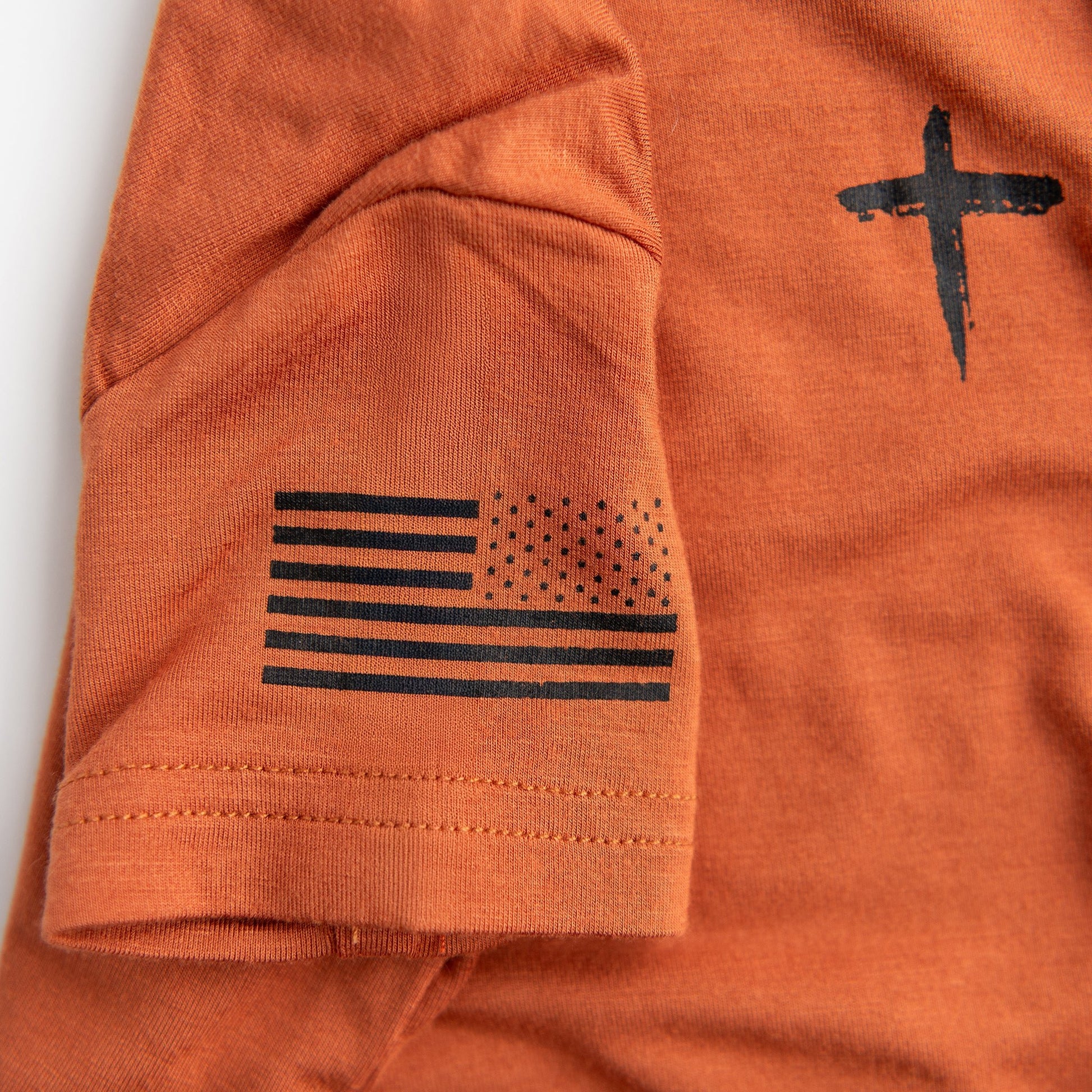 A close-up detail of the sleeve on the Apostle Living Inc. burnt orange onesie, highlighting the distressed black American flag and part of the cross design.