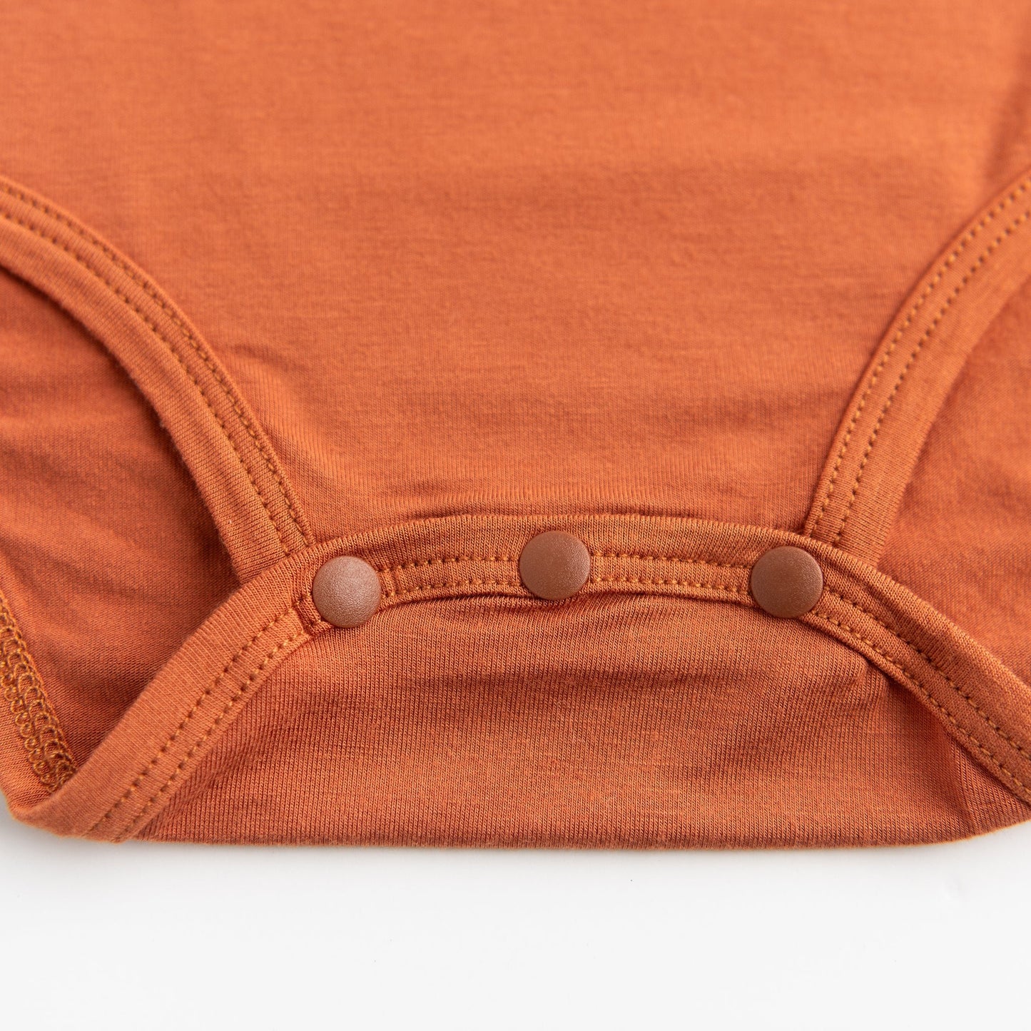 A close-up detail shot of the three-snap closure at the bottom of the Apostle Living Inc. Faith and Freedom baby onesie in burnt orange.