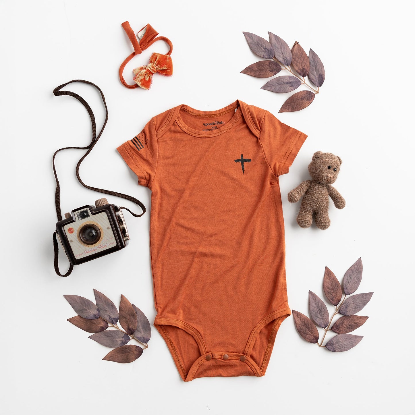 Apostle Living Inc. Faith and Freedom baby onesie in burnt orange, featuring a black cross and American flag, styled with a teddy bear, vintage camera, and leaves.