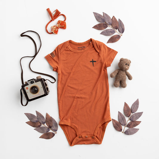 Apostle Living Inc. Faith and Freedom baby onesie in burnt orange, featuring a black cross and American flag, styled with a teddy bear, vintage camera, and leaves.