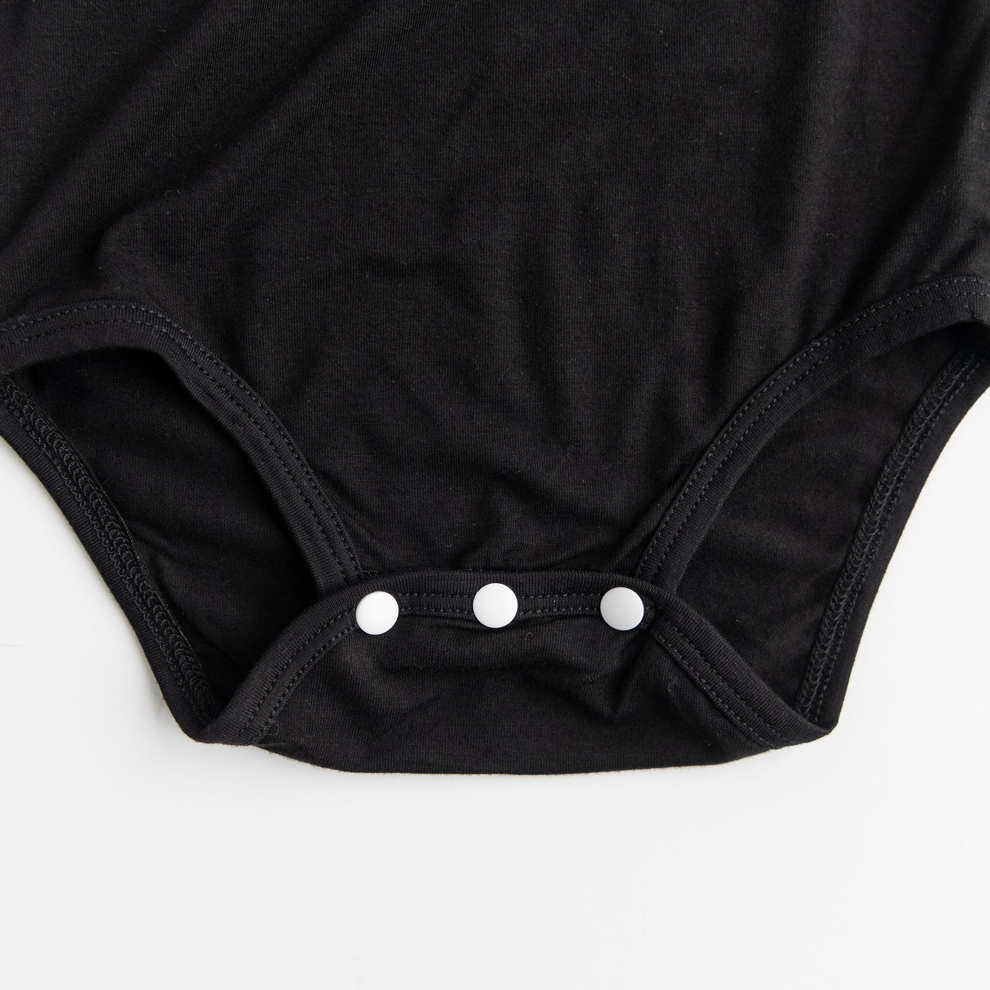 
A close-up detail shot of the three-snap closure on the bottom of the Apostle Living Inc. Faith and Freedom baby onesie in black, showing the three white snaps.