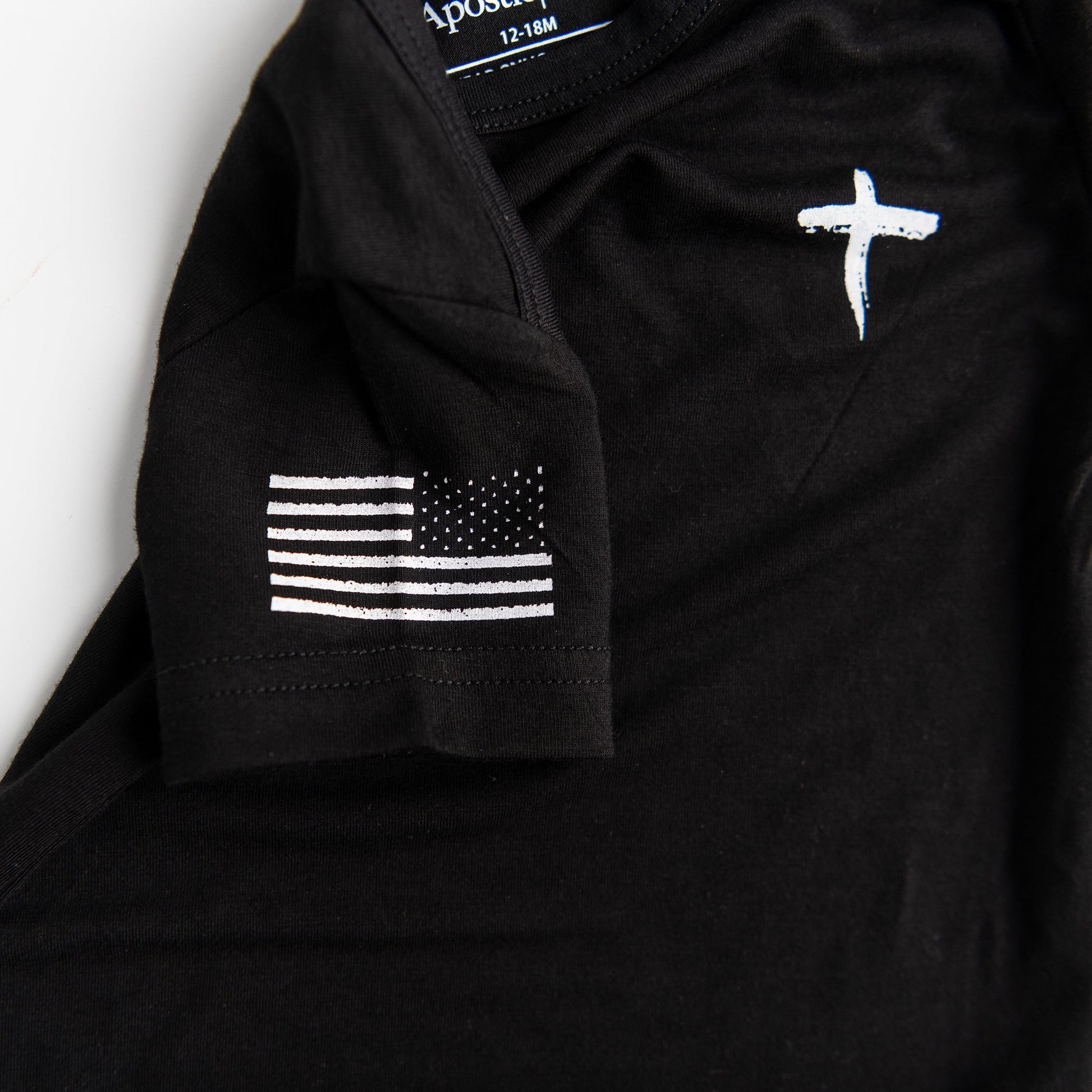 A close-up detail shot of the black Apostle Living Inc. Faith and Freedom onesie, highlighting the distressed white cross on the chest and the distressed white American flag on the sleeve.