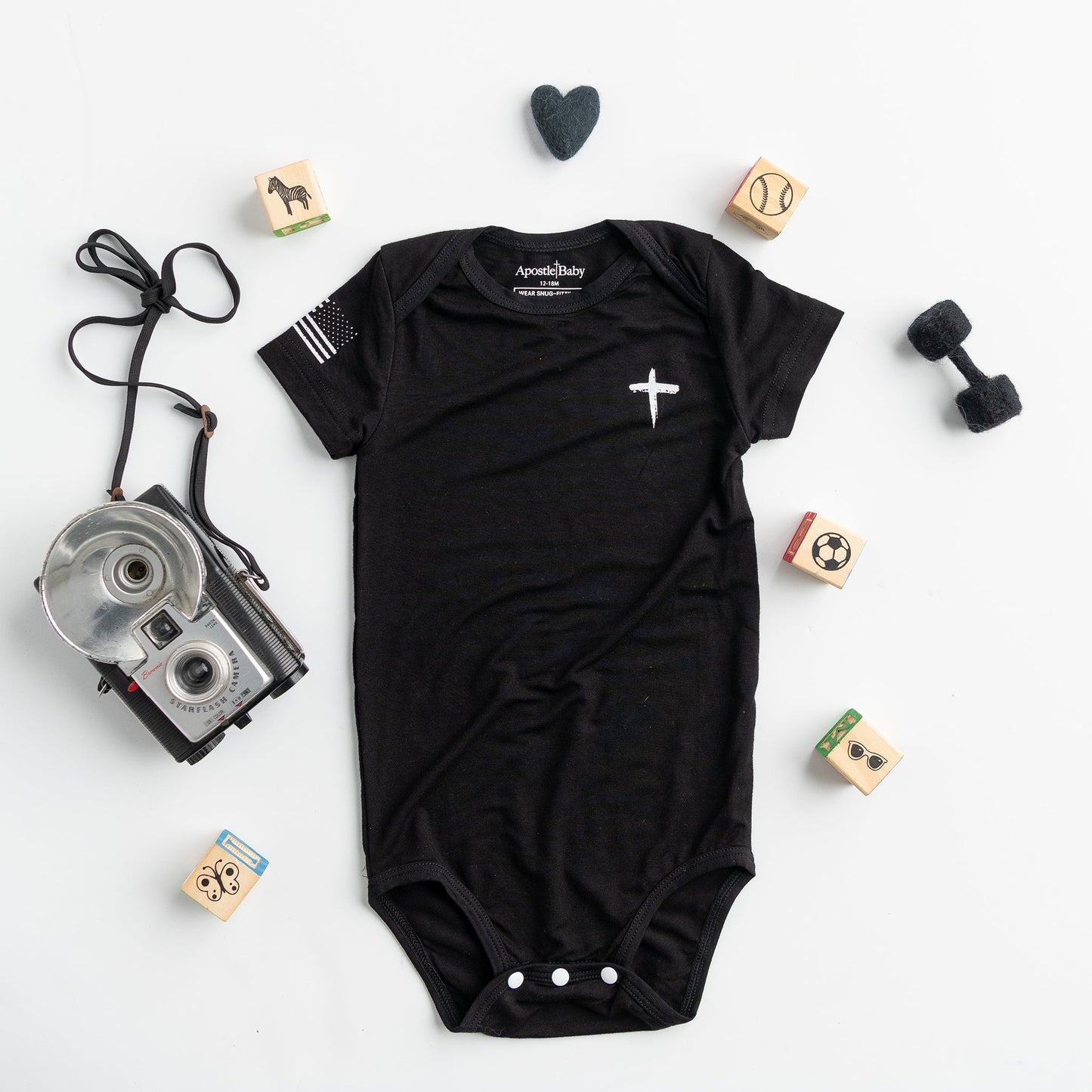 Black baby onesie with a white cross and American flag on the sleeve by Apostle Living Inc., part of the Faith and Freedom collection.