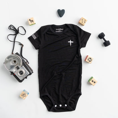 Black baby onesie with a white cross and American flag on the sleeve by Apostle Living Inc., part of the Faith and Freedom collection.