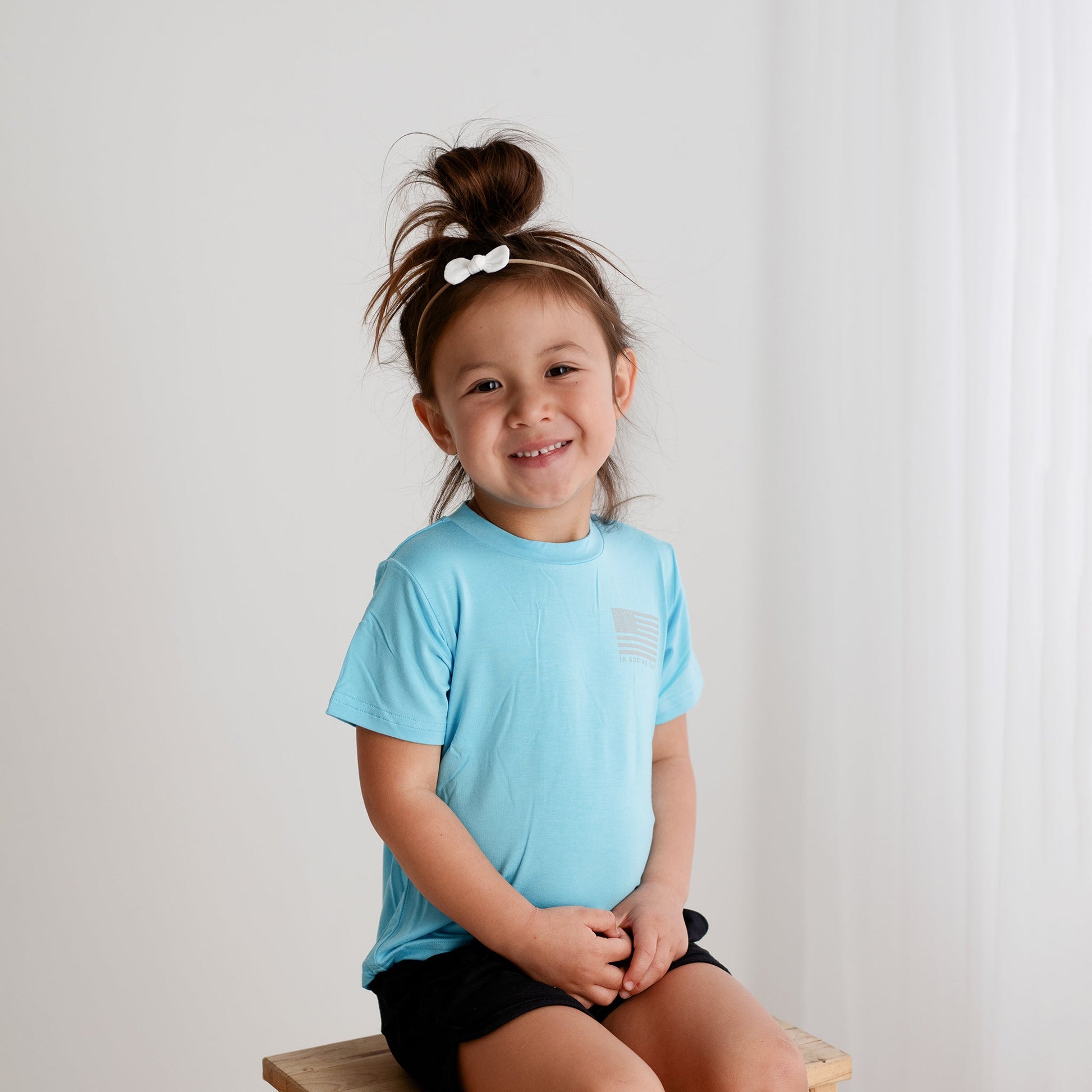 A young girl from Apostle Living smiles while sitting on a wooden stool, wearing a light blue patriotic tee with a small distressed U.S. flag graphic and the text 'IN GOD WE TRUST' on the chest.