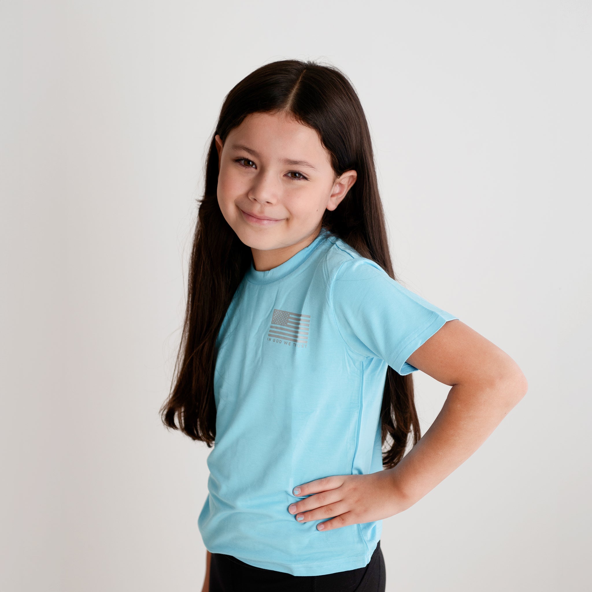 A young girl from Apostle Living models a light blue patriotic tee with a small distressed U.S. flag graphic and the text 'IN GOD WE TRUST' on the chest.