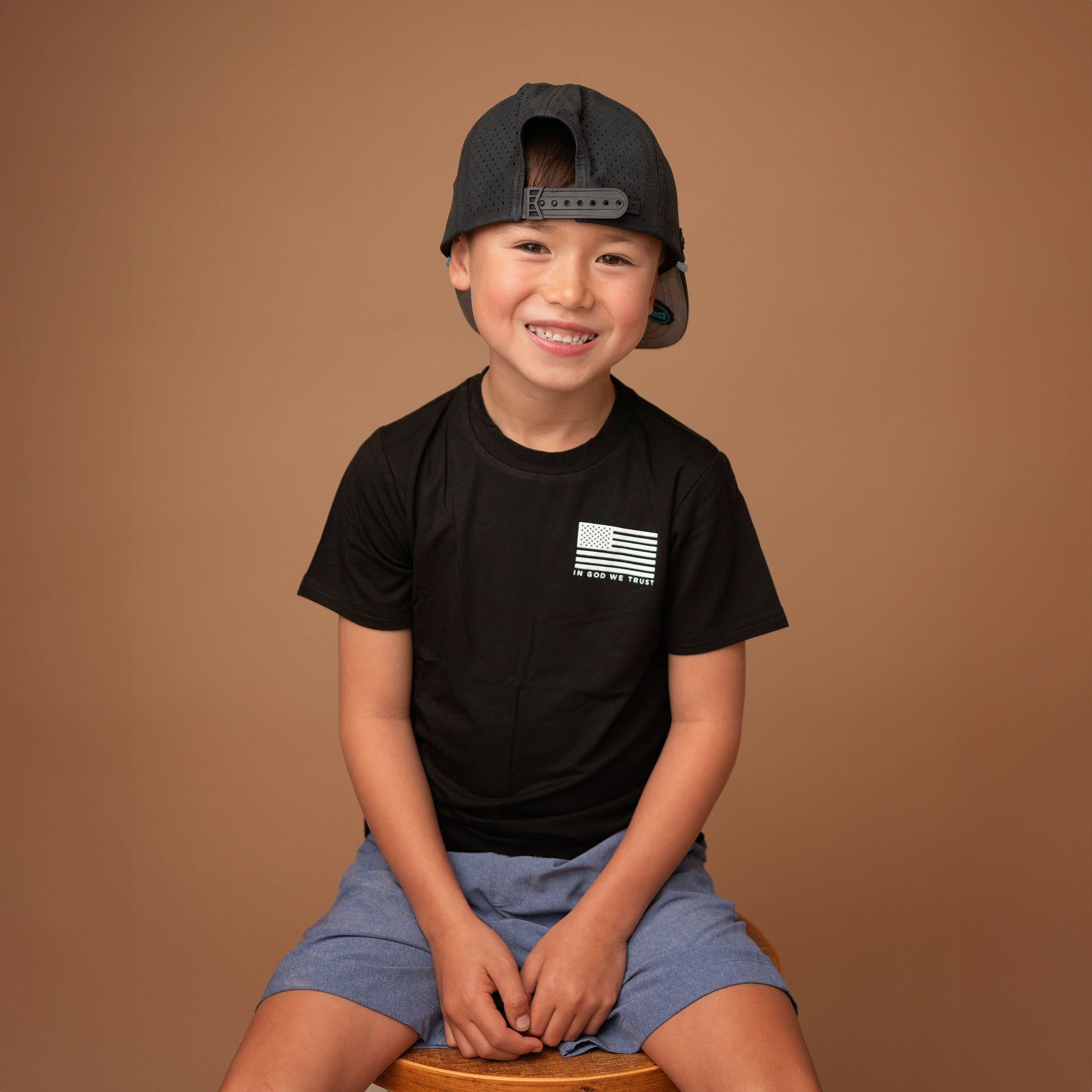 A young boy from Apostle Living sits barefoot on a wooden stool, smiling and wearing a black patriotic tee with a small distressed U.S. flag graphic and the text 'IN GOD WE TRUST' on the chest.