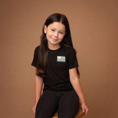 `A young girl from Apostle Living sits on a wooden stool, wearing a black patriotic tee with a white distressed USA.