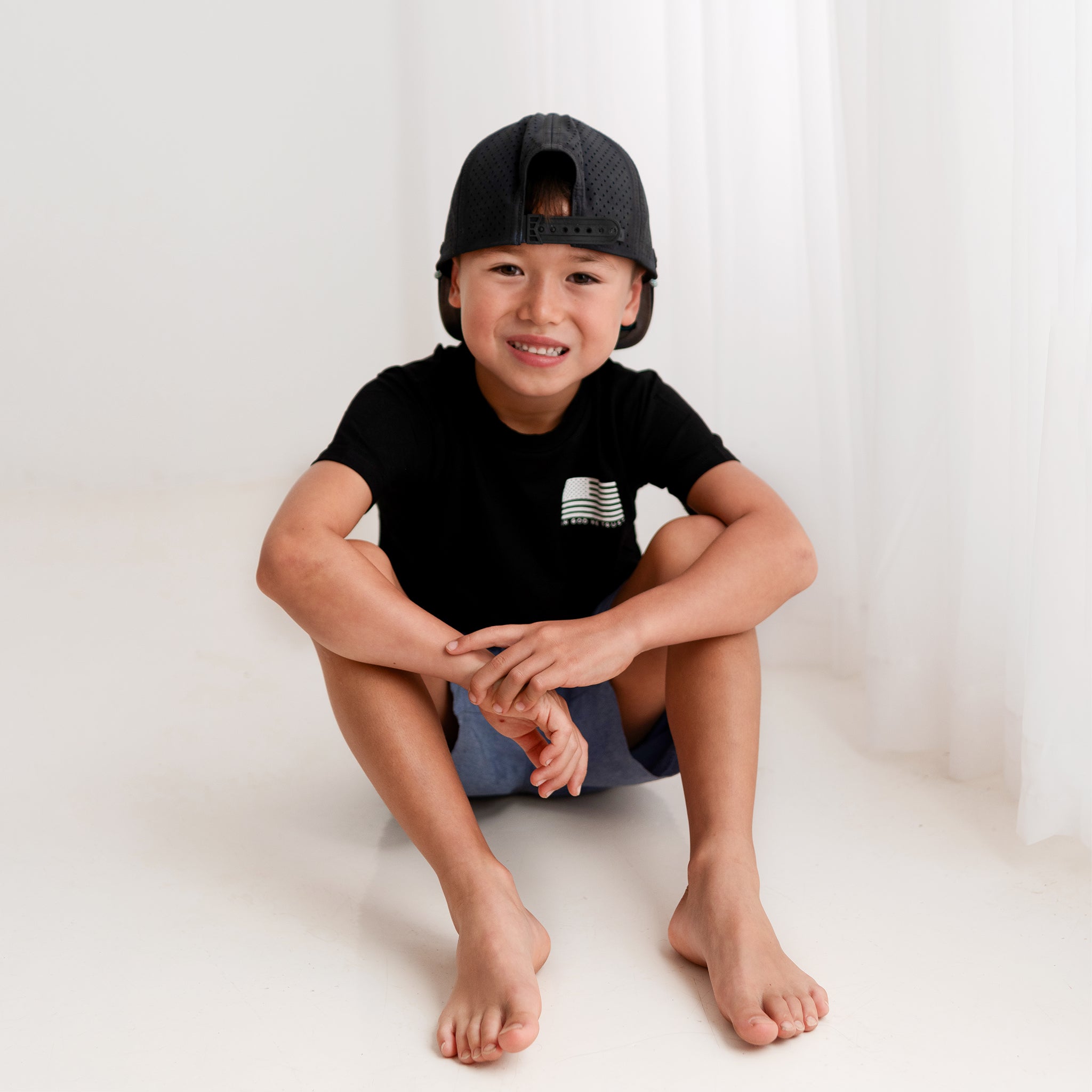 A young boy from Apostle Living smiles while sitting on a white floor, wearing a black patriotic bamboo tee with a U.S. flag and the text 'IN GOD WE TRUST' on the left chest.