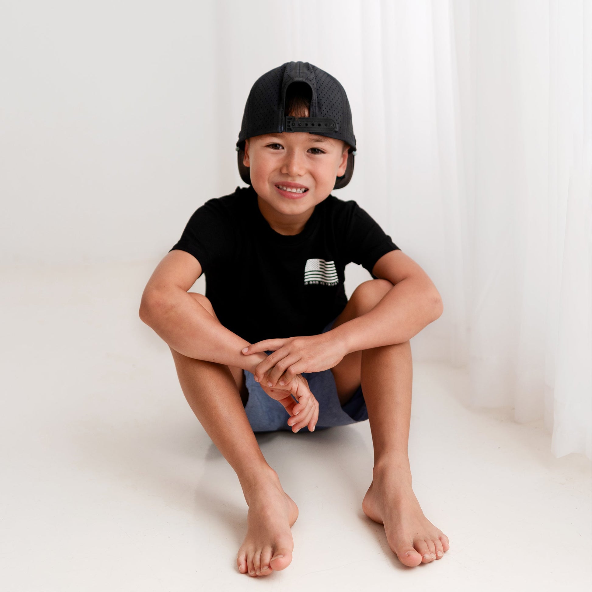 A young boy from Apostle Living smiles while sitting on a white floor, wearing a black patriotic bamboo tee with a U.S. flag and the text 'IN GOD WE TRUST' on the left chest.