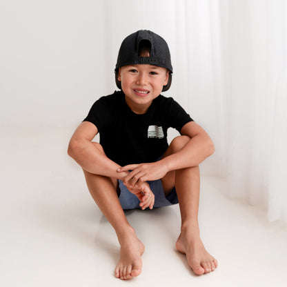 A young boy from Apostle Living smiles while sitting on a white floor, wearing a black patriotic bamboo tee with a U.S. flag and the text 'IN GOD WE TRUST' on the left chest.