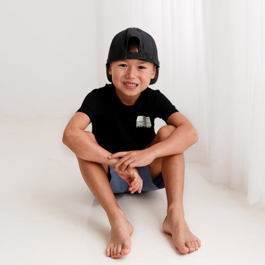 A young boy from Apostle Living smiles while sitting on a white floor, wearing a black patriotic bamboo tee with a U.S. flag and the text 'IN GOD WE TRUST' on the left chest.