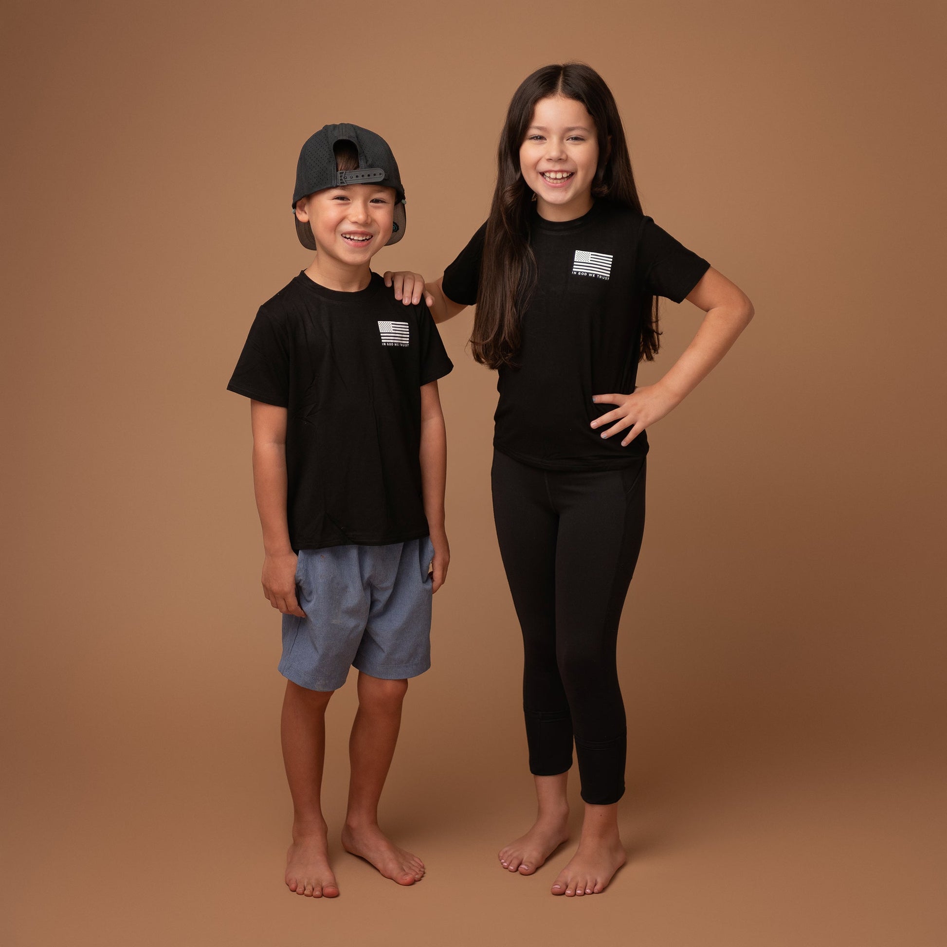 A boy and girl from Apostle Living stand together, both wearing black patriotic tees with a distressed U.S. flag graphic and the text 'IN GOD WE TRUST' on the chest.