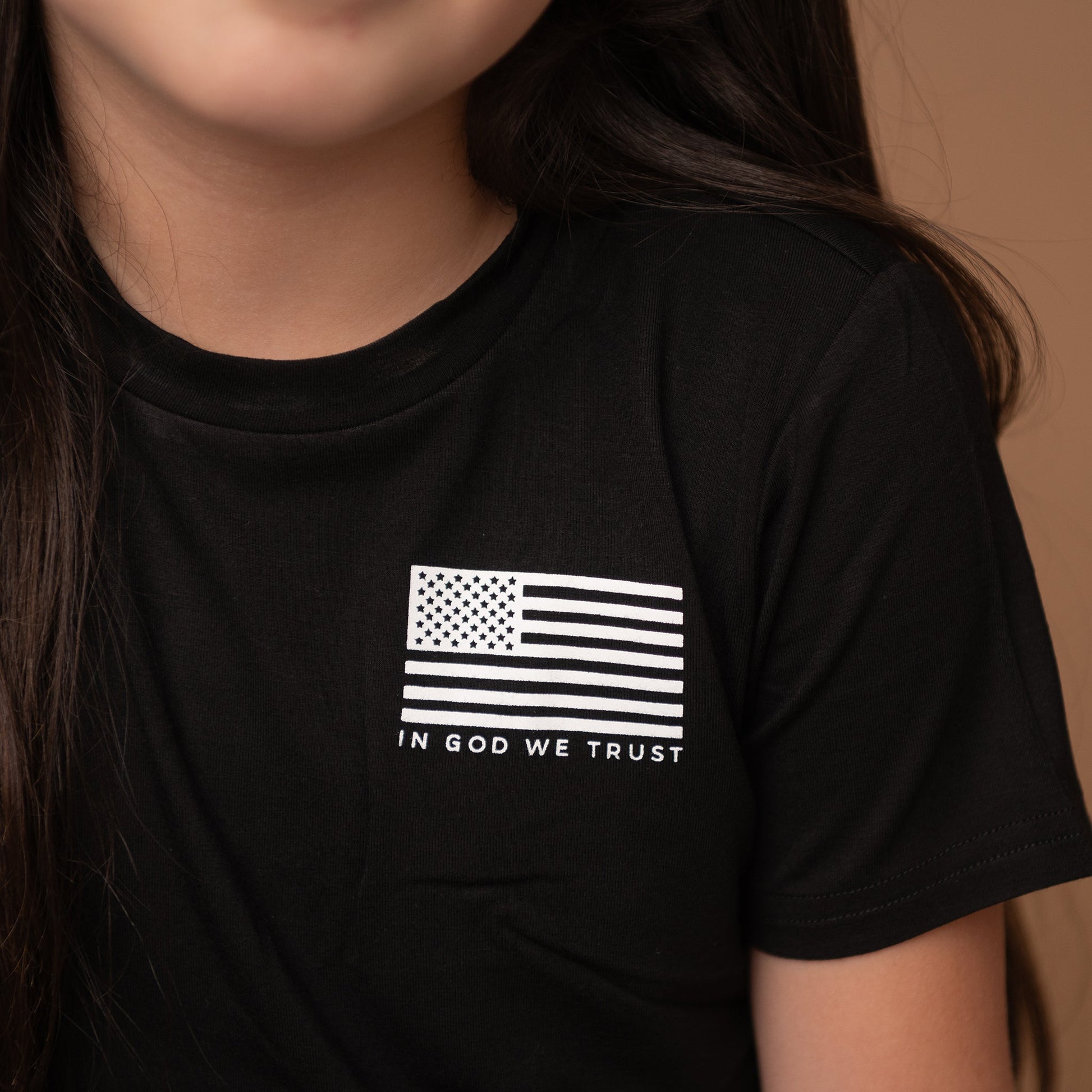 A close-up product shot of a black t-shirt, focusing on a white graphic of a distressed U.S. flag and the text 'IN GOD WE TRUST' on the chest.