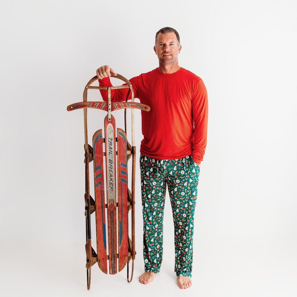 A man smiling and holding a vintage red sled while wearing a red long-sleeve shirt and Apostle Living Inc. green Christmas pajama pants with a festive pattern.