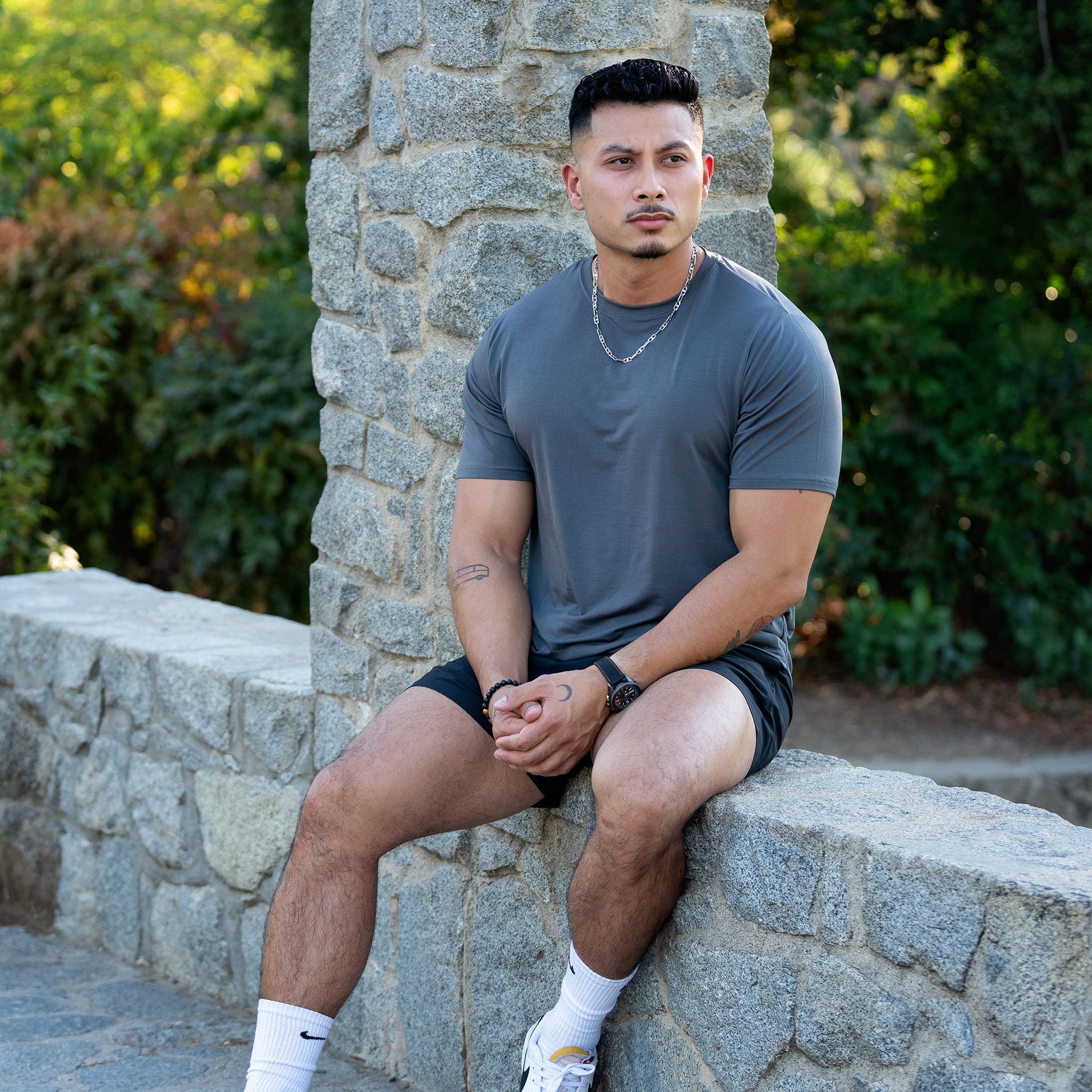 A man wearing a gray lyocell tee and black shorts, sitting on an outdoor stone wall.