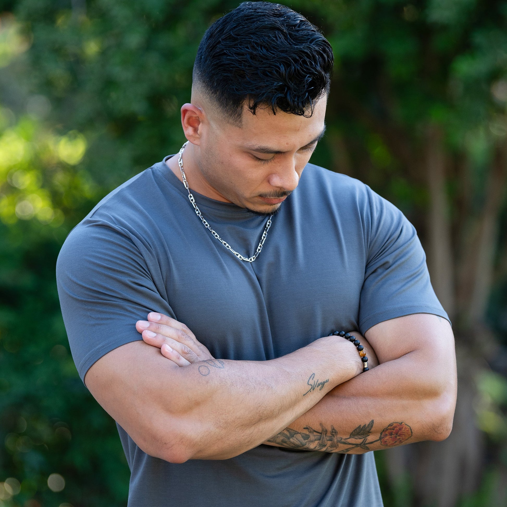 A man modeling a gray lyocell tee, with his arms crossed and looking down in a close-up outdoor shot.