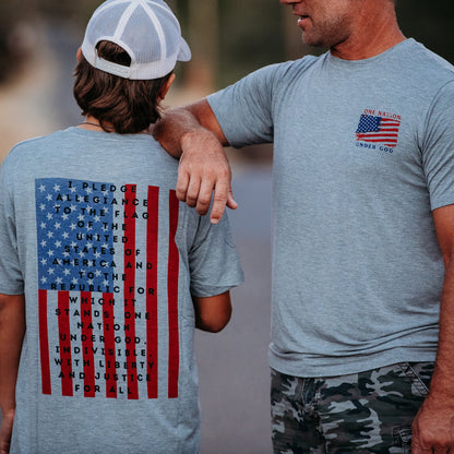 A father and son from Apostle Living stand together, wearing gray patriotic tees with a U.S. flag graphic. The shirt features the Pledge of Allegiance, and has a smaller flag and the text 'ONE NATION UNDER GOD'.
