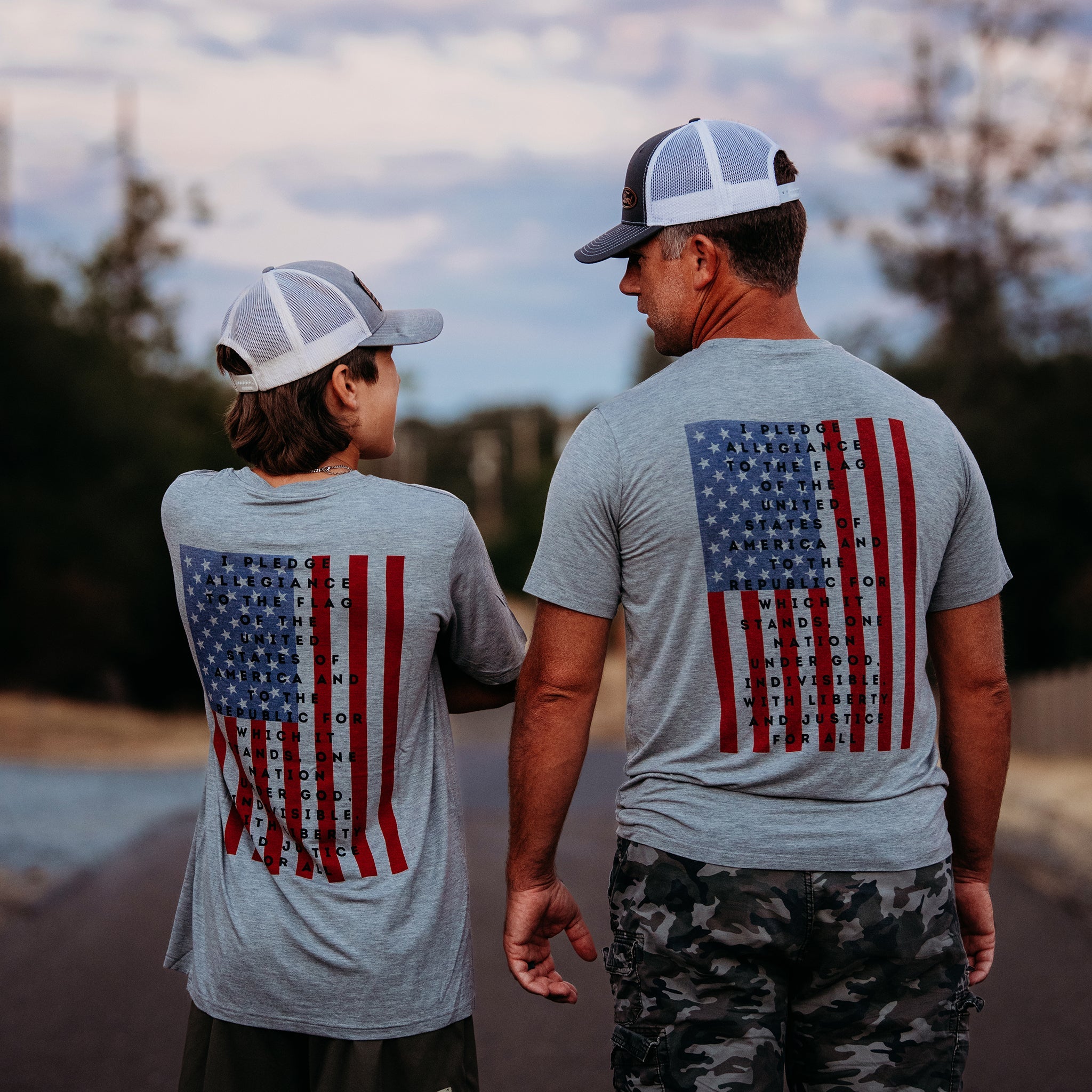 A father and son from Apostle Living, viewed from behind, wearing matching gray patriotic tees featuring the U.S. flag and the Pledge of Allegiance.