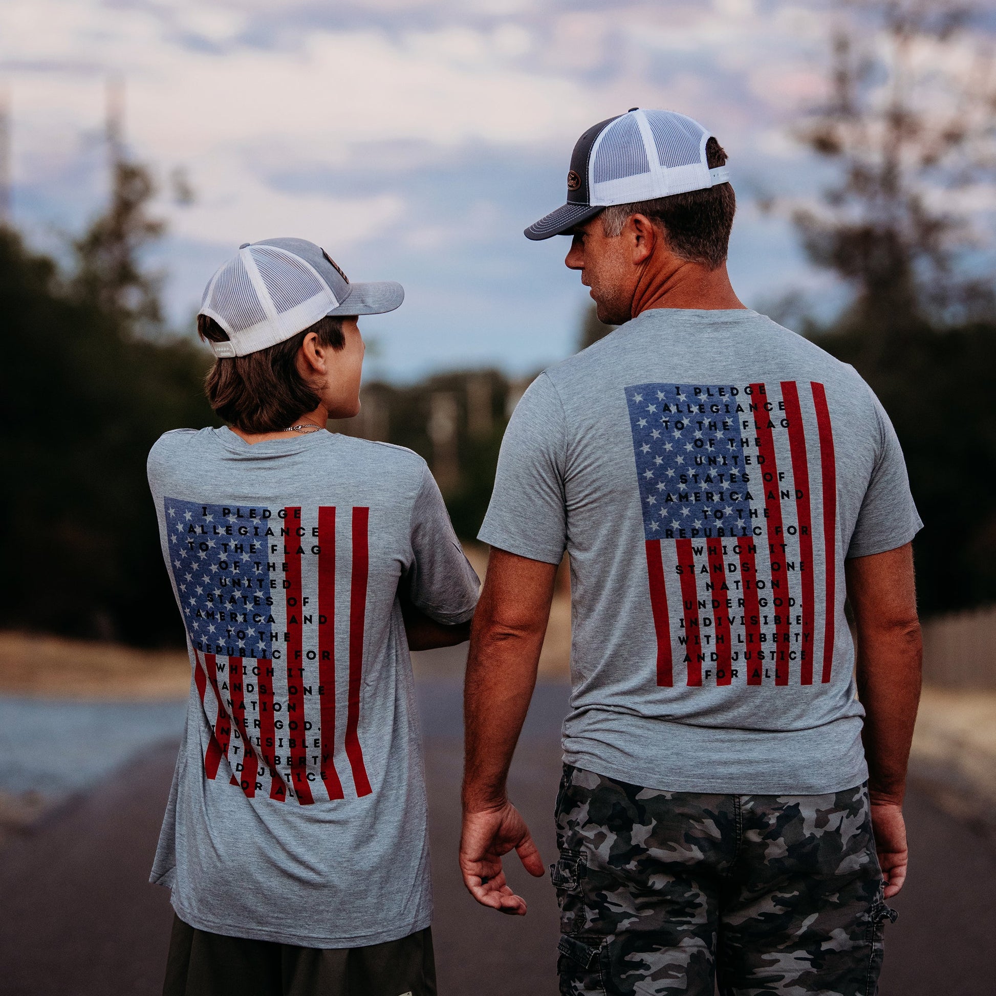 A father and son from Apostle Living, viewed from behind, wearing matching gray patriotic tees featuring the U.S. flag and the Pledge of Allegiance.