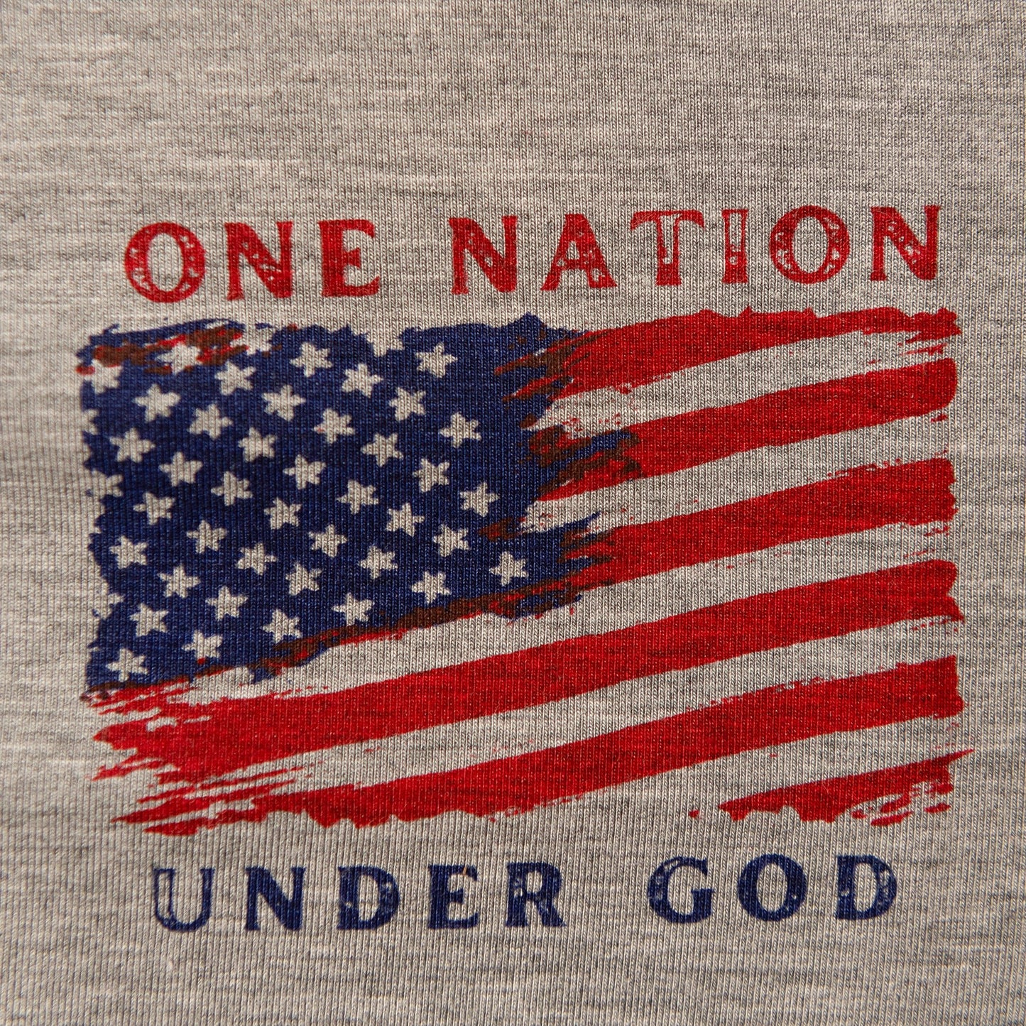 A close-up product shot of a gray t-shirt with a distressed patriotic graphic featuring a U.S. flag and the text 'ONE NATION UNDER GOD'.