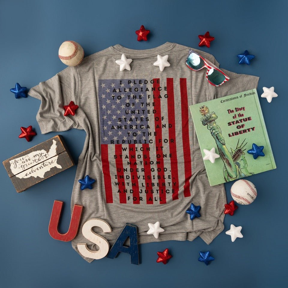 A flat-lay product shot of a gray t-shirt from Apostle Living featuring the Pledge of Allegiance printed over a U.S. flag graphic, styled with patriotic items like a book on the Statue of Liberty, baseballs, and wooden USA letters.